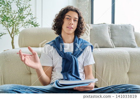 Curly teenage student boy gesturing and holding notebook during online class. Curly teenage student boy gesturing and holding notebook during online class. 141033819