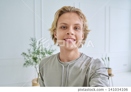 Happy teenage boy smiling at camera while taking selfie in bright modern room Happy teenage boy smiling at camera while taking selfie in bright modern room 141034106