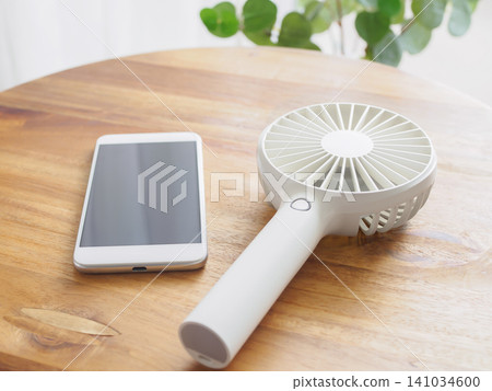桌上放著一把手持風扇和一支智慧型手機。 桌上放著一把手持風扇和一支智慧型手機。 141034600