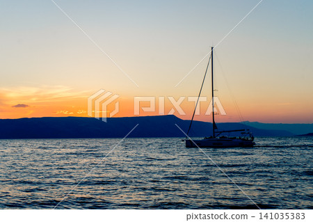 Elegant sailboat silhouetted against a glowing sunset over calm sea with distant mountains, creating a peaceful summer travel and sailing scene. Elegant sailboat silhouetted against a glowing sunset over calm sea with distant mountains, creating a peaceful summer travel and sailing scene. 141035383