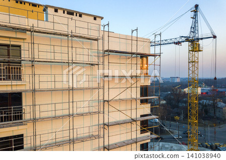 Apartment or office tall unfinished building under construction. Brick wall in scaffolding, shiny windows and tower crane on urban landscape and blue sky background. Drone aerial photography. Apartment or office tall unfinished building under construction. Brick wall in scaffolding, shiny windows and tower crane on urban landscape and blue sky background. Drone aerial photography. 141038940