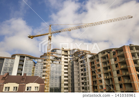 Apartment or office tall building under construction. Brick walls, glass windows, scaffolding and concrete support pillars. Tower crane on bright blue sky copy space background. Apartment or office tall building under construction. Brick walls, glass windows, scaffolding and concrete support pillars. Tower crane on bright blue sky copy space background. 141038961