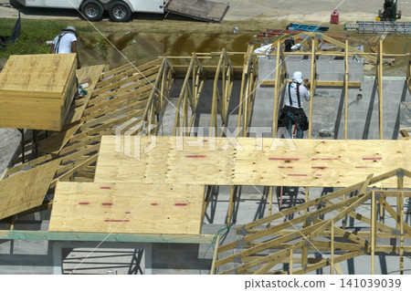 Aerial view of builders working on unfinished residential house with wooden roof frame structure under construction in Florida suburban area Aerial view of builders working on unfinished residential house with wooden roof frame structure under construction in Florida suburban area 141039039