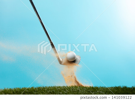 Golf ball explode from Tee off. Golfer hit ball with metal iron club to sand explosion on green. Golf club hit ball on green tee off with sand flying scatter. Blue Sky background isolated Golf ball explode from Tee off. Golfer hit ball with metal iron club to sand explosion on green. Golf club hit ball on green tee off with sand flying scatter. Blue Sky background isolated 141039122