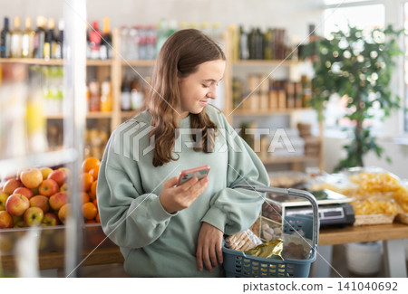 Visiting store, teen girl with phone is looking at something on shelf of showcase. Visiting store, teen girl with phone is looking at something on shelf of showcase. 141040692