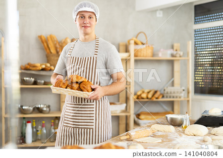 Portrait of young happy male baker working in bakehouse, holding tray with fresh bakery goods on kitchen Portrait of young happy male baker working in bakehouse, holding tray with fresh bakery goods on kitchen 141040804