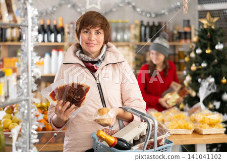Cheerful woman with shopping basket in shop holding and choosing dried meat or jamon for festive table for Christmas with daughter looking goods on food shelves in background Cheerful woman with shopping basket in shop holding and choosing dried meat or jamon for festive table for Christmas with daughter looking goods on food shelves in background 141041102