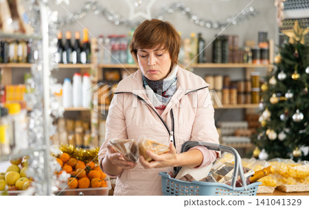 Portrait of adult female buyer with shopping cart or basket holding assortment of cheese choosing for christmas table in grocery shop Portrait of adult female buyer with shopping cart or basket holding assortment of cheese choosing for christmas table in grocery shop 141041329