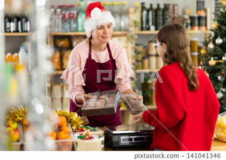 Friendly woman shop seller in festive hat posing behind counter with Christmas goods and offering cookies to girl Friendly woman shop seller in festive hat posing behind counter with Christmas goods and offering cookies to girl 141041346