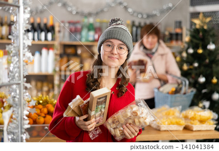 Portrait of young girl choosing sweets in supermarket, mother choosing groceries to festive table in background Portrait of young girl choosing sweets in supermarket, mother choosing groceries to festive table in background 141041384