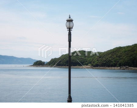 一盞孤零零的路燈矗立在大海和島嶼的輪廓映襯下;初夏的八代海,一幅令人懷舊的景象。 一盞孤零零的路燈矗立在大海和島嶼的輪廓映襯下;初夏的八代海,一幅令人懷舊的景象。 141041385