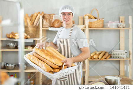European young bakery male seller stands proudly in front of fresh bread holding basket of freshly baked baguettes in kitchen European young bakery male seller stands proudly in front of fresh bread holding basket of freshly baked baguettes in kitchen 141041451