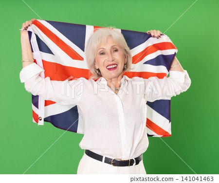 Positive senior woman in white suit holds big national flag of United Kingdom, green background 141041463