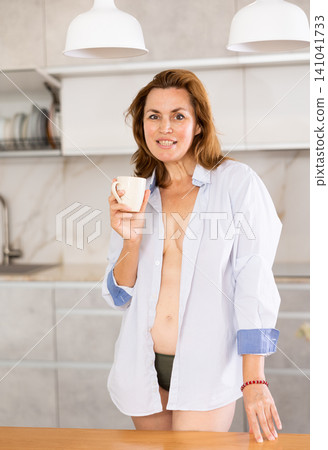 Charming topless woman in shirt with coffee mug at kitchen Charming topless woman in shirt with coffee mug at kitchen 141041733