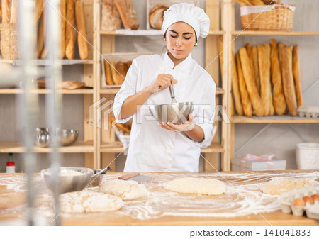 Young female baker whisking dough in bowl Young female baker whisking dough in bowl 141041833