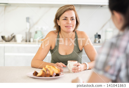Smiling middle-aged woman talking friendly while drinking tea in the kitchen Smiling middle-aged woman talking friendly while drinking tea in the kitchen 141041863