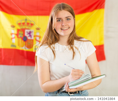 Cheerful smiling teen girl, student in casual wear writing in spiral notepad against of national flag of Spain Cheerful smiling teen girl, student in casual wear writing in spiral notepad against of national flag of Spain 141041864