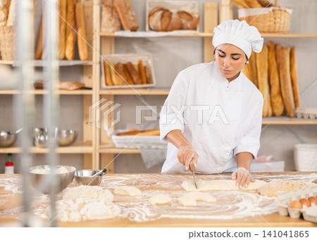 Young female baker cutting dough on table in bakehouse Young female baker cutting dough on table in bakehouse 141041865