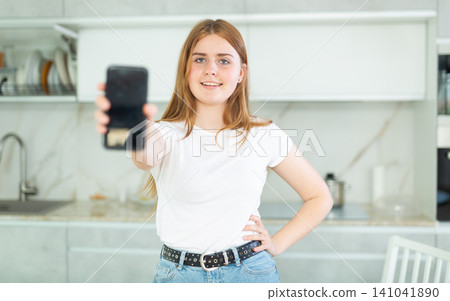 Portrait of positive teen girl showing modern mobile phone with black screen in cozy kitchen. Gadget with empty free space for mock up Portrait of positive teen girl showing modern mobile phone with black screen in cozy kitchen. Gadget with empty free space for mock up 141041890