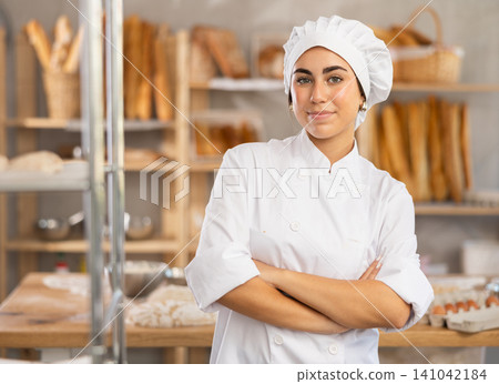 Young female baker stands in bake house, ready to greet customers Young female baker stands in bake house, ready to greet customers 141042184