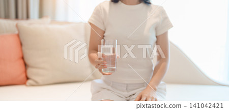 Healthy woman hold glass of clean mineral water glass on sofa at home, female drink pure water for hydration and wellness, fresh skin care, daily routine water intake, Refreshment and lifestyle 141042421