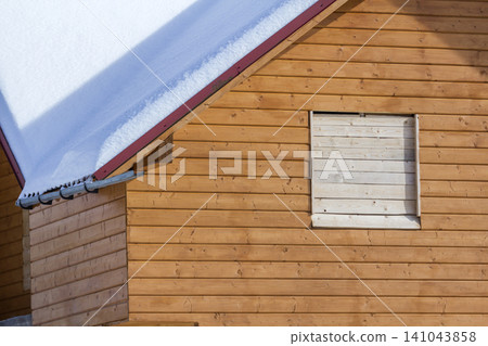 Detail of new wooden ecological traditional cottage of natural lumber materials with steep roof covered with snow on sunny winter day. Old traditions and modern professional construction concept. Detail of new wooden ecological traditional cottage of natural lumber materials with steep roof covered with snow on sunny winter day. Old traditions and modern professional construction concept. 141043858