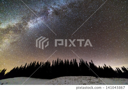 Winter mountains night landscape panorama. Milky Way bright constellation in dark blue starry sky over dark spruce pine trees forest, soft glow on horizon after sunset. Wide angle shot. Winter mountains night landscape panorama. Milky Way bright constellation in dark blue starry sky over dark spruce pine trees forest, soft glow on horizon after sunset. Wide angle shot. 141043867