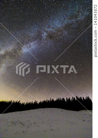 Winter mountains night landscape panorama. Milky Way bright constellation in dark blue starry sky over dark spruce pine trees forest, soft glow on horizon after sunset. Wide angle shot. Winter mountains night landscape panorama. Milky Way bright constellation in dark blue starry sky over dark spruce pine trees forest, soft glow on horizon after sunset. Wide angle shot. 141043872