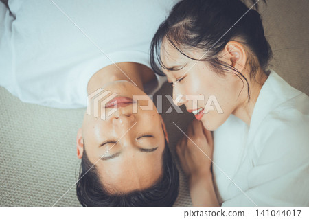 A couple portrait from above, depicting a girlfriend smiling as she peers over her sleeping boyfriend on a rug, showcasing her sleep beauty image. A couple portrait from above, depicting a girlfriend smiling as she peers over her sleeping boyfriend on a rug, showcasing her sleep beauty image. 141044017