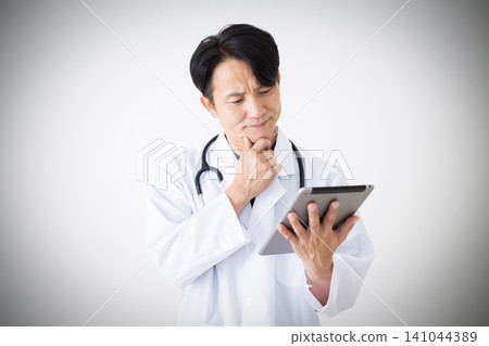 A middle-aged male doctor in a white coat is shown looking troubled while holding a tablet. 141044389