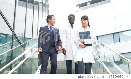 一位商人和一群穿著白大褂的跨國工程師一邊走下樓梯一邊交談 一位商人和一群穿著白大褂的跨國工程師一邊走下樓梯一邊交談 141044644