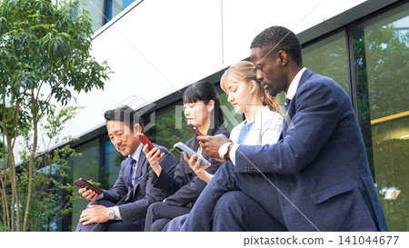 A group of multinational business people sitting on an outdoor bench and looking at their smartphones A group of multinational business people sitting on an outdoor bench and looking at their smartphones 141044677