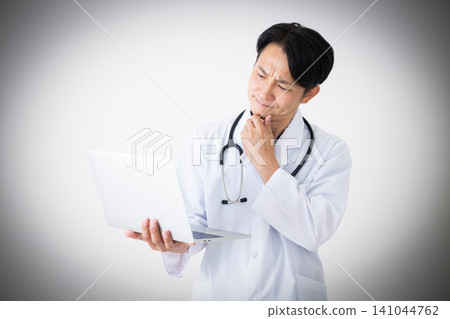 A middle-aged male doctor in a white coat, holding a laptop and deep in thought. A middle-aged male doctor in a white coat, holding a laptop and deep in thought. 141044762