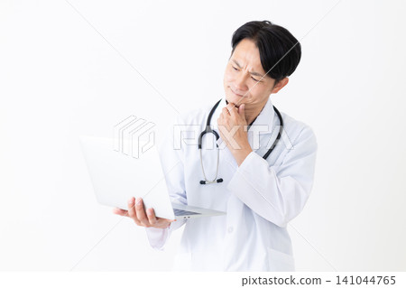 A middle-aged male doctor in a white coat, holding a laptop and deep in thought. A middle-aged male doctor in a white coat, holding a laptop and deep in thought. 141044765