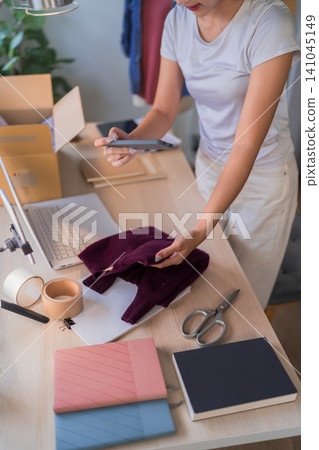 Young Asian woman online merchant scanning clothing with smartphone while organizing inventory at workspace with laptop and packages Young Asian woman online merchant scanning clothing with smartphone while organizing inventory at workspace with laptop and packages 141045149