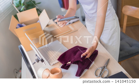 Young Asian woman online merchant scanning package with smartphone while preparing orders with laptop and shipping boxes at home Young Asian woman online merchant scanning package with smartphone while preparing orders with laptop and shipping boxes at home 141045150