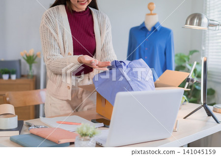 Asian woman small business owner packing blue shirt order in bright home office studio workspace Asian woman small business owner packing blue shirt order in bright home office studio workspace 141045159