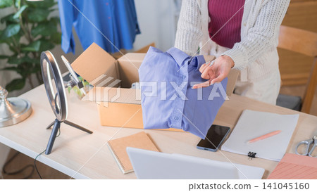 Asian woman online merchant packing blue shirt into cardboard box while preparing order shipment at home workspace Asian woman online merchant packing blue shirt into cardboard box while preparing order shipment at home workspace 141045160