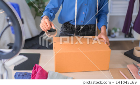 Online merchant in blue shirt sealing cardboard box with packing tape for ecommerce order shipping at home office Online merchant in blue shirt sealing cardboard box with packing tape for ecommerce order shipping at home office 141045169