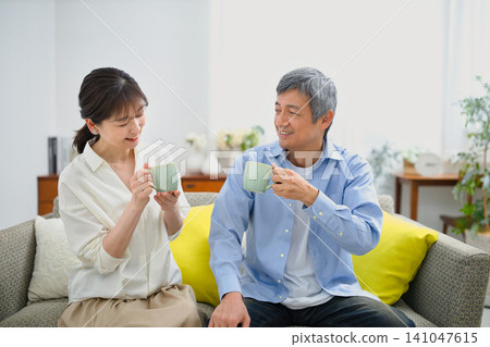 A smiling couple in their 60s sitting on a sofa and drinking coffee. A smiling couple in their 60s sitting on a sofa and drinking coffee. 141047615
