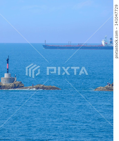 一座燈塔矗立在漂浮於美麗夏日大海中的岩石岬角上,人們在岸邊享受釣魚的樂趣,地平線上還有一艘貨輪。 一座燈塔矗立在漂浮於美麗夏日大海中的岩石岬角上,人們在岸邊享受釣魚的樂趣,地平線上還有一艘貨輪。 141047729
