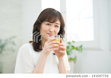 A woman in her 60s drinking vegetable juice in her living room. A woman in her 60s drinking vegetable juice in her living room. 141047738