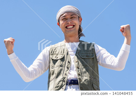 A man in work clothes strikes a victory pose against a blue sky. A man in work clothes strikes a victory pose against a blue sky. 141048455