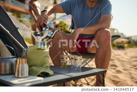Man pouring tea on a sandy beach during a relaxed summer vacation Man pouring tea on a sandy beach during a relaxed summer vacation 141048517
