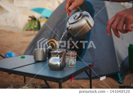 Person pouring tea from a kettle into a teapot at a beach campsite Person pouring tea from a kettle into a teapot at a beach campsite 141048520
