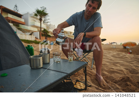 Man pouring tea while camping on a sandy beach at sunset with tent and coastal homes Man pouring tea while camping on a sandy beach at sunset with tent and coastal homes 141048521