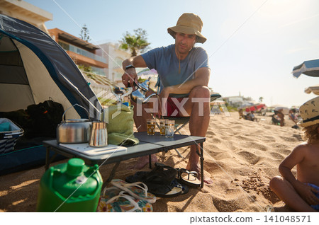 Man pouring tea at a beach campsite with child and tent on a sunny summer day Man pouring tea at a beach campsite with child and tent on a sunny summer day 141048571