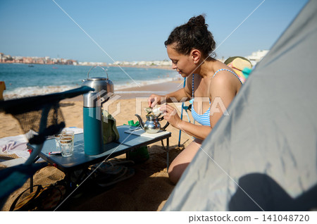 Woman preparing tea at a beach picnic by the sea on a sunny summer day Woman preparing tea at a beach picnic by the sea on a sunny summer day 141048720