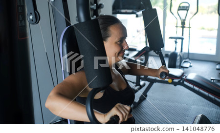 Young woman in black sportswear performing chest press exercise on a machine in a modern equipped gym. Upper body training concept Young woman in black sportswear performing chest press exercise on a machine in a modern equipped gym. Upper body training concept 141048776