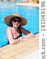 Woman in sunglasses and wide-brimmed hat swimming leisurely in a sunny pool 141049196